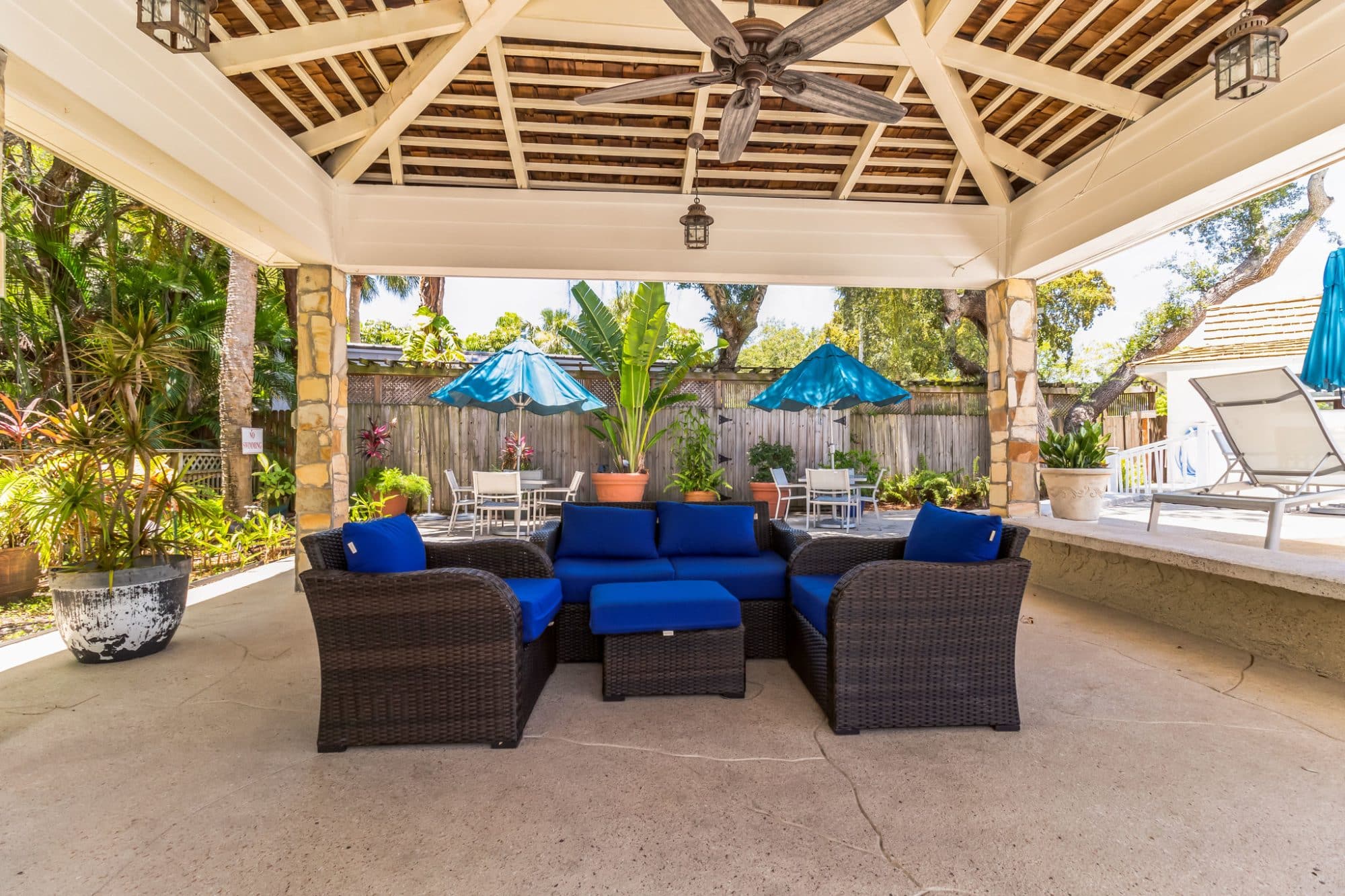 Outdoor lounge under a covered patio with wicker furniture and blue cushions, surrounded by tropical plants and dining tables with blue umbrellas in naples fl